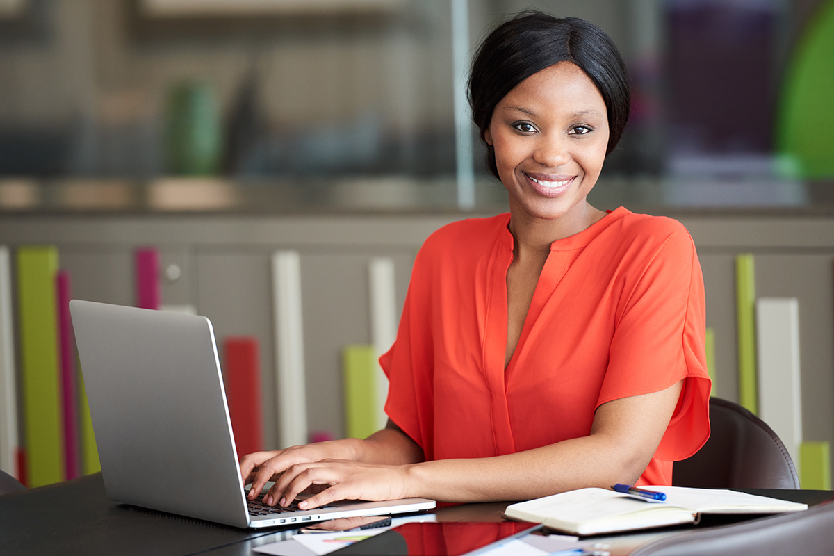 Beautiful well dressed young black businesswoman looking at the camera while sitting at her desk in her colourful office environment, busy typing on her modern laptop computer.